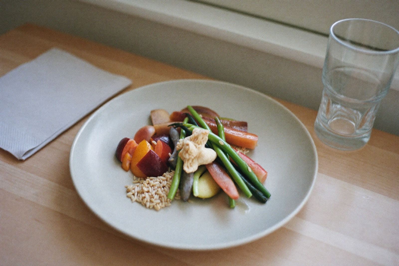 Un plato con verduras, granos y fruta sobre una mesa de madera clara, junto a un vaso de agua