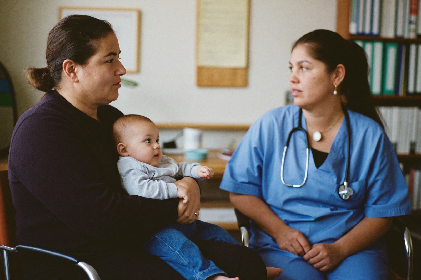 Una mamá con su bebé en brazos conversando con una profesional de la salud en un consultorio luminoso