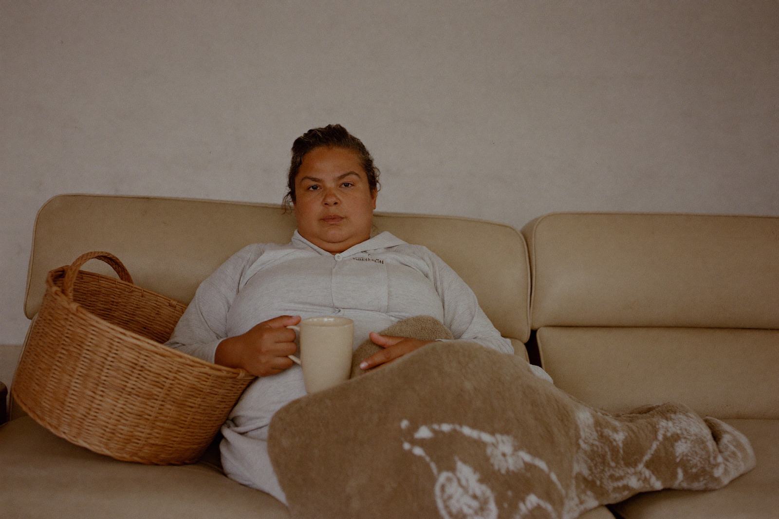 Una mujer con una manta sobre los hombros sostiene a su bebé recostada en un sillón, con una taza en la mesa lateral