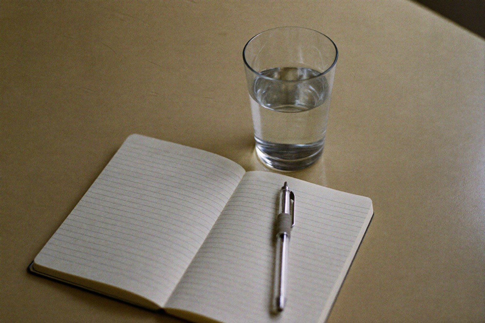 Un vaso de agua sobre una mesa de madera junto a una libreta abierta y un bolígrafo, con luz de ventana