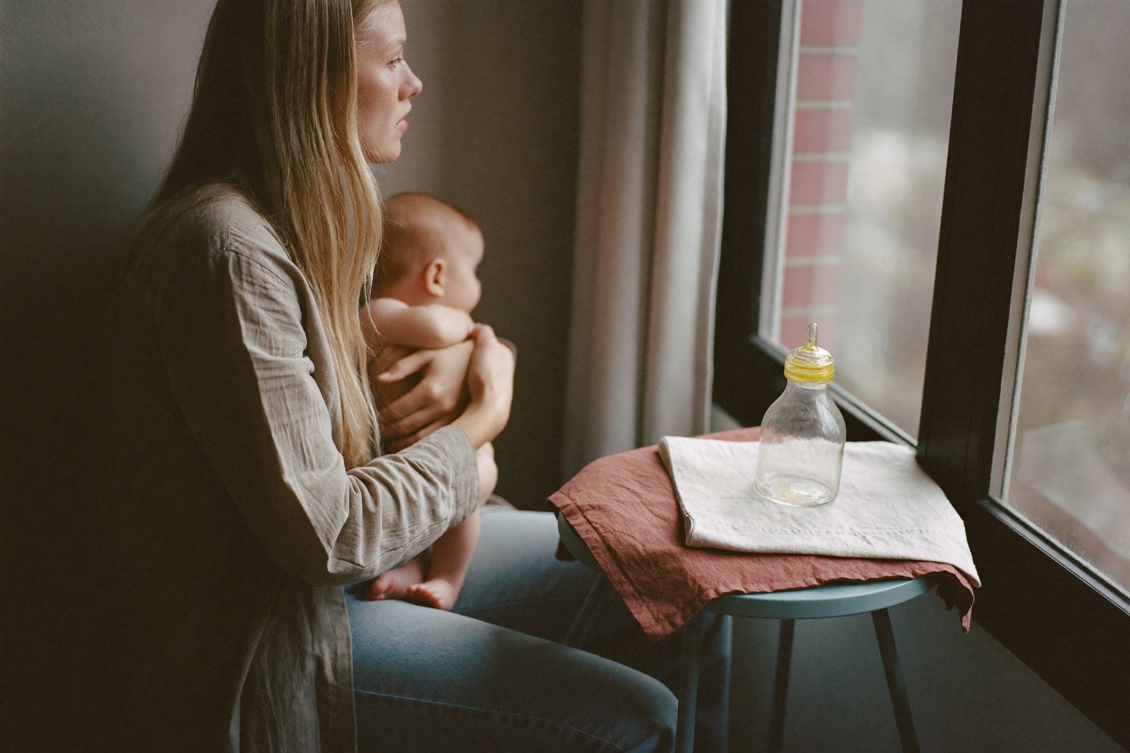 Una madre sentada junto a una ventana sostiene a su bebé en brazos, con un biberón limpio y una muselina doblada sobre la mesa lateral