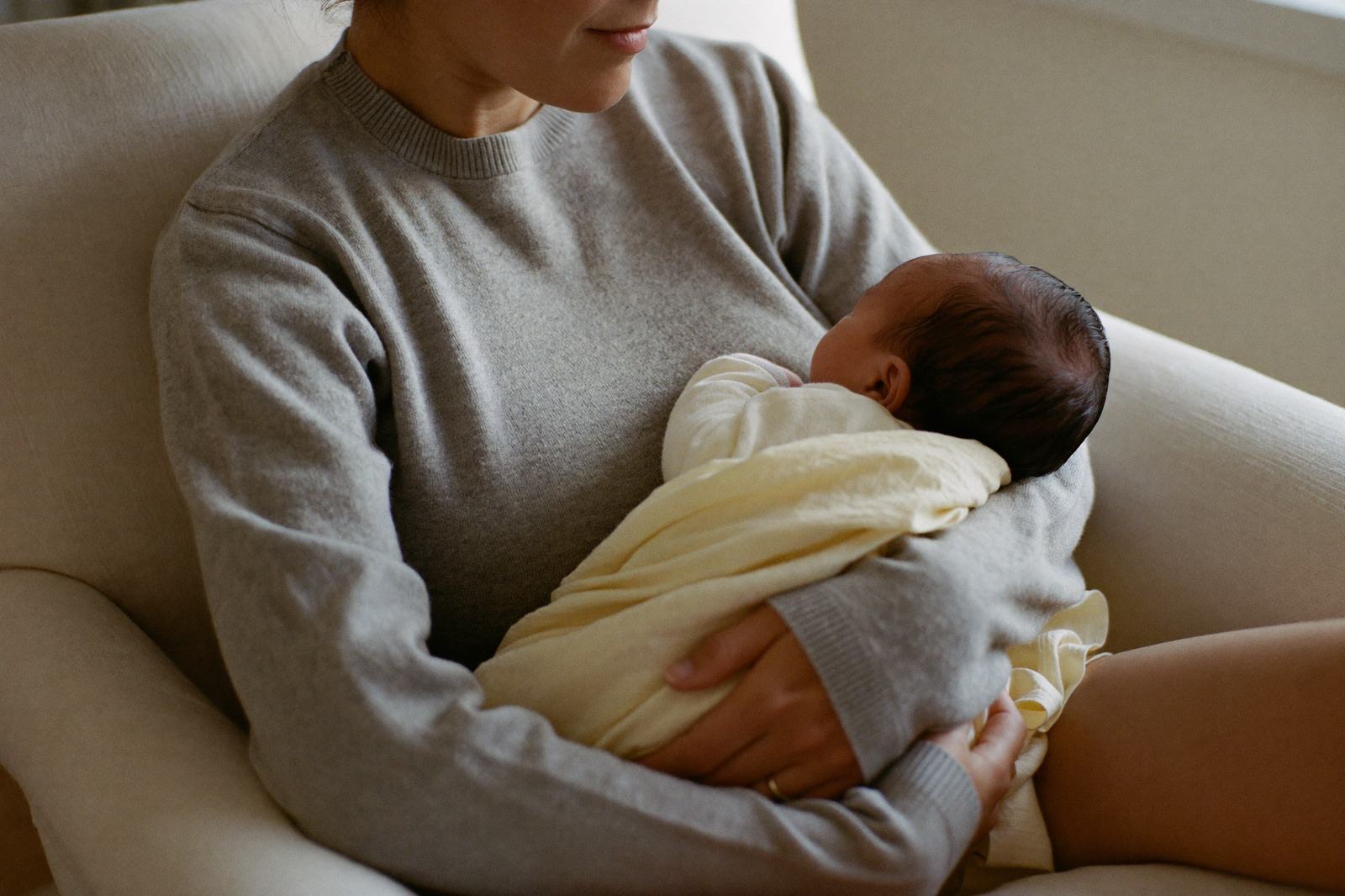 Una mamá sentada en un sillón amamanta a su bebé en posición de cuna, con la cabeza del bebé apoyada en el pliegue del codo