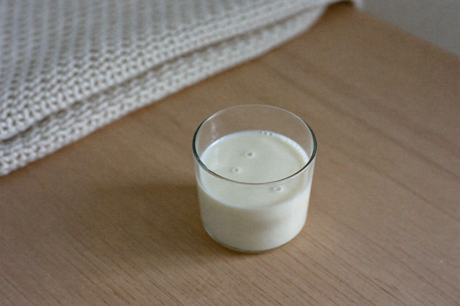 Vaso de vidrio con leche materna extraída sobre una mesa de madera clara, junto a una manta doblada