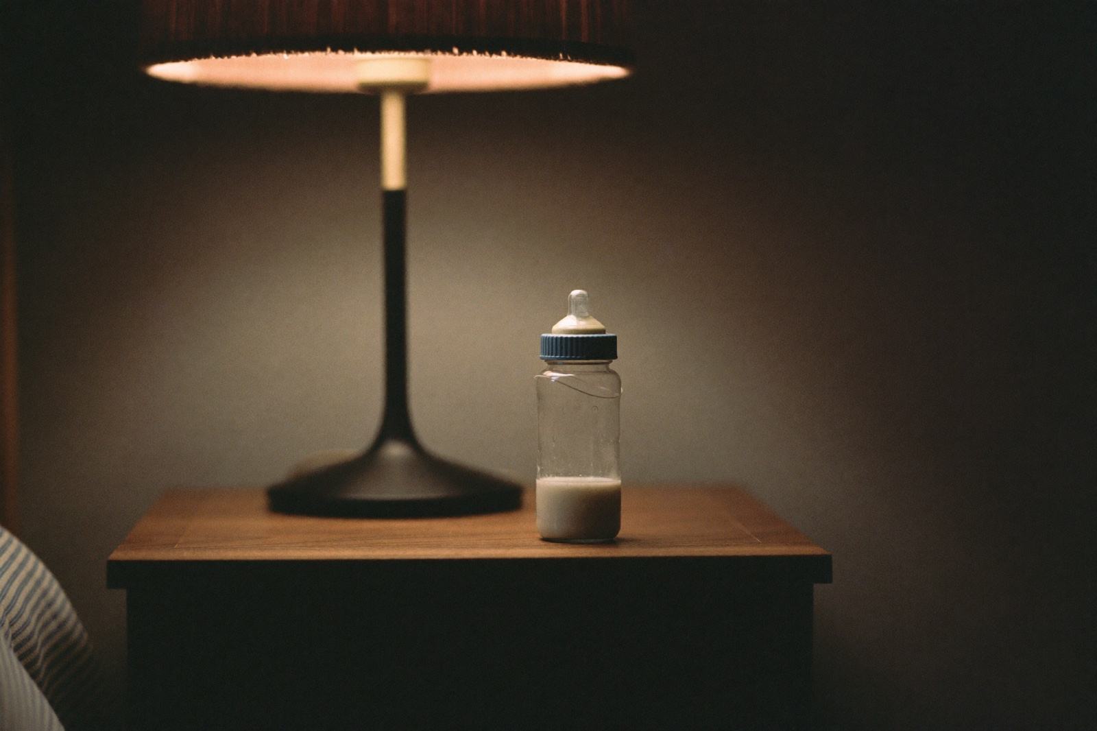 Un biberón a medio tomar sobre una mesa de noche de madera, con una lámpara de luz cálida encendida detrás