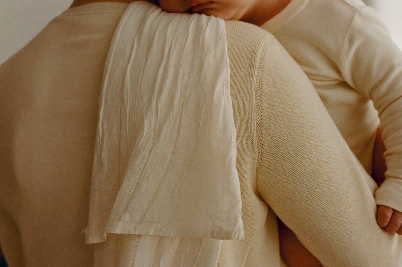 Bebé recostado sobre el hombro de un adulto, con una tela de muselina sobre la ropa, en una habitación de luz cálida