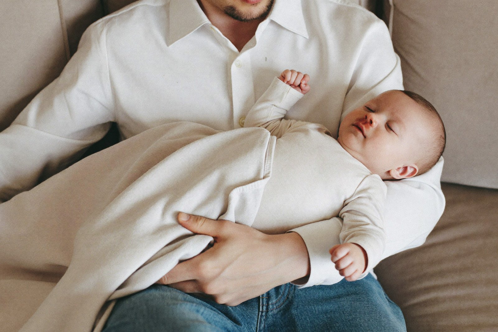 Un bebé dormido en brazos de su papá, envuelto en una manta ligera, en un sofá