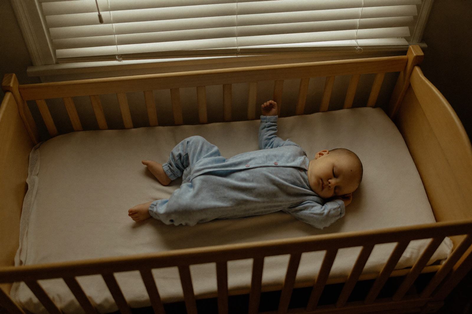 Un bebé dormido de lado en su cuna de madera con una manta ligera, en una habitación con la persiana entrecerrada