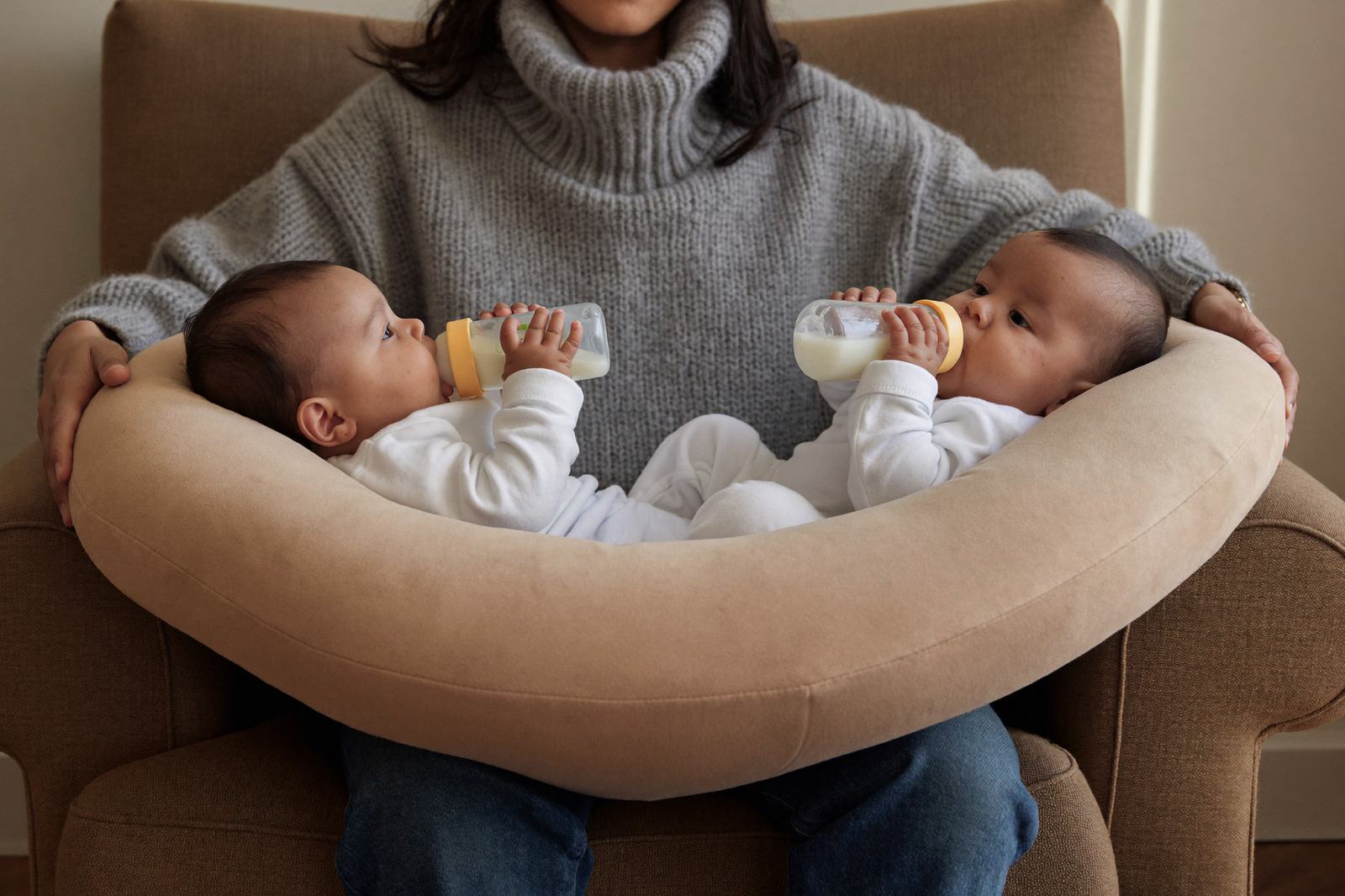 Mamá sentada en un sillón dando de comer a dos bebés a la vez con una almohada de lactancia