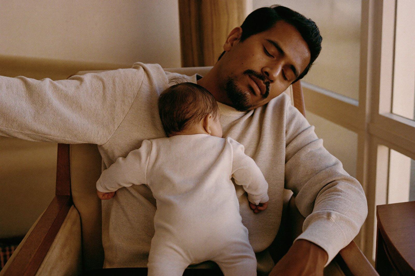 Un bebé dormido contra el hombro de su papá, que también se quedó dormido en un sillón junto a la ventana, con luz de tarde