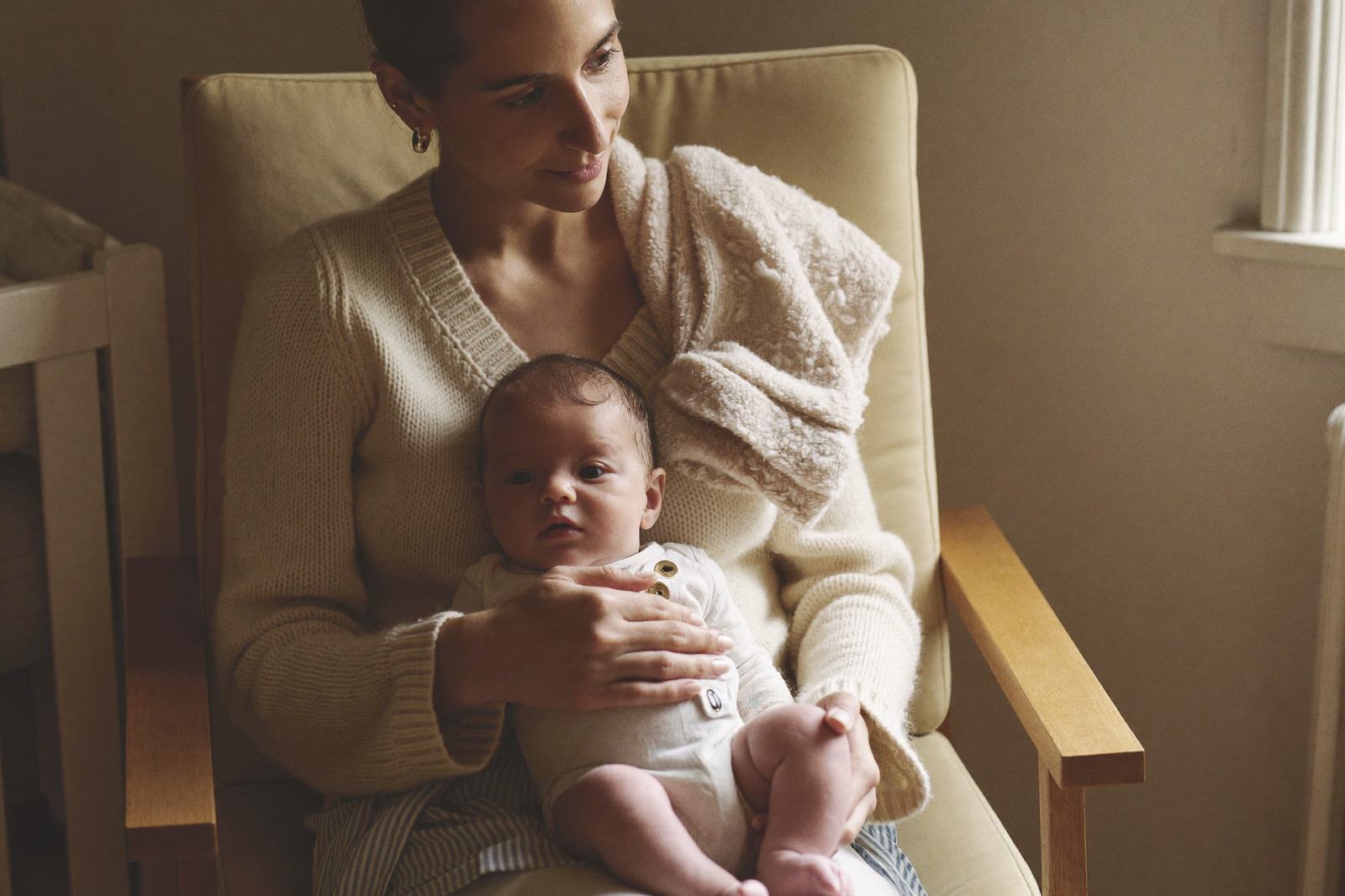 Mamá sentada en un sillón junto a la cuna con su bebé en brazos, con luz de ventana