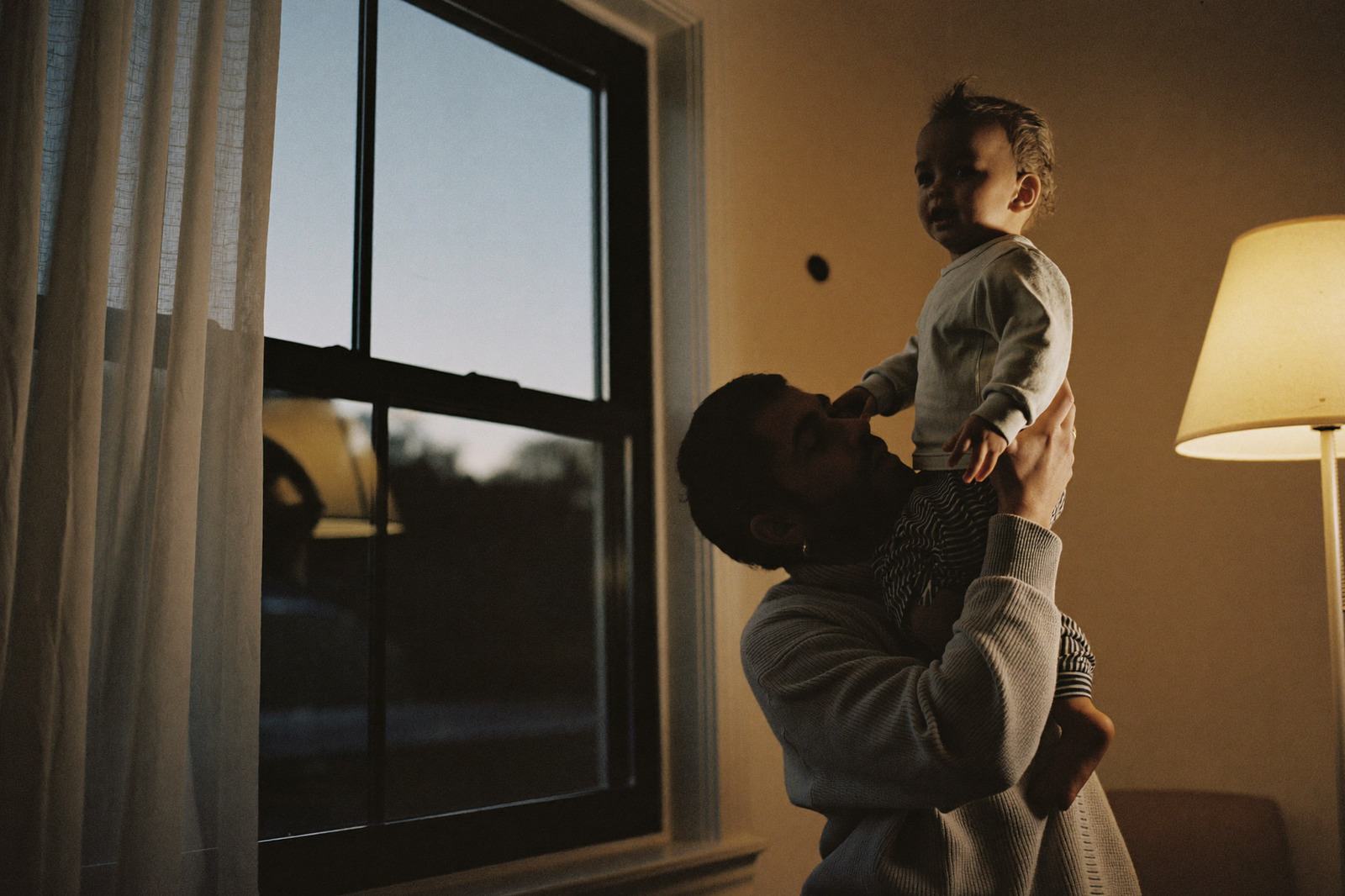 Papá cargando en alto a su hijo pequeño junto a una ventana al anochecer, con una lámpara encendida