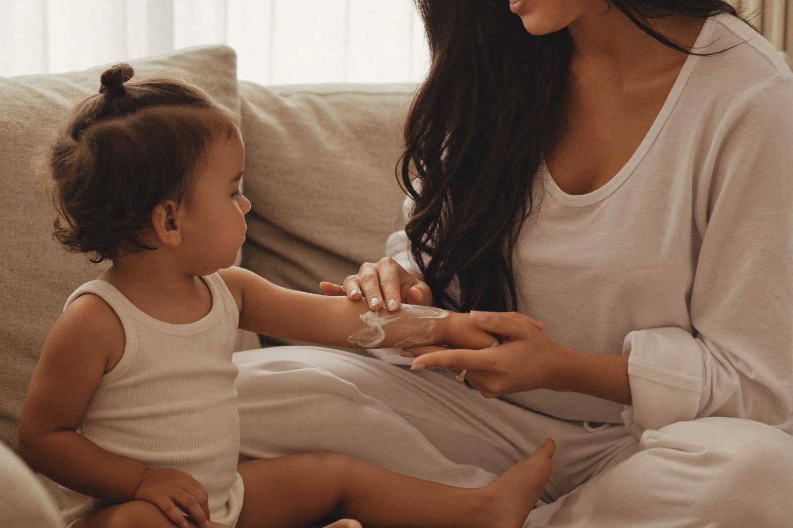 Mamá aplicando crema en el brazo de su hija pequeña, sentadas en un sillón con luz de día