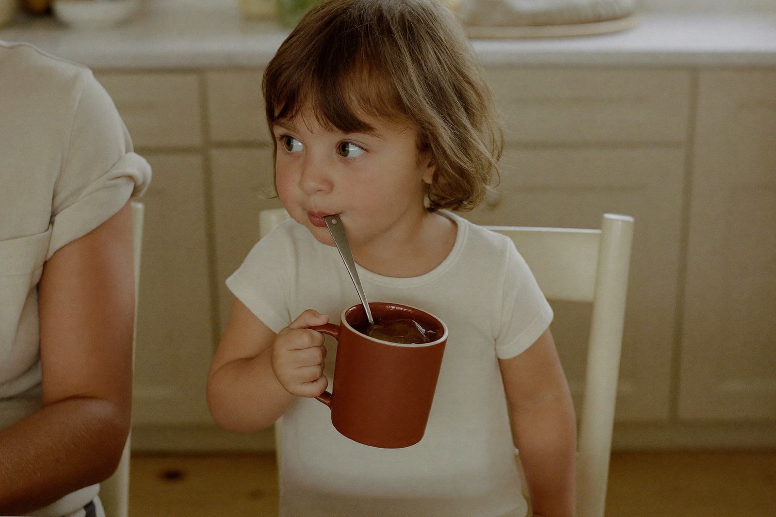 Niño pequeño tomando de una taza en la mesa de la cocina, con un adulto a su lado