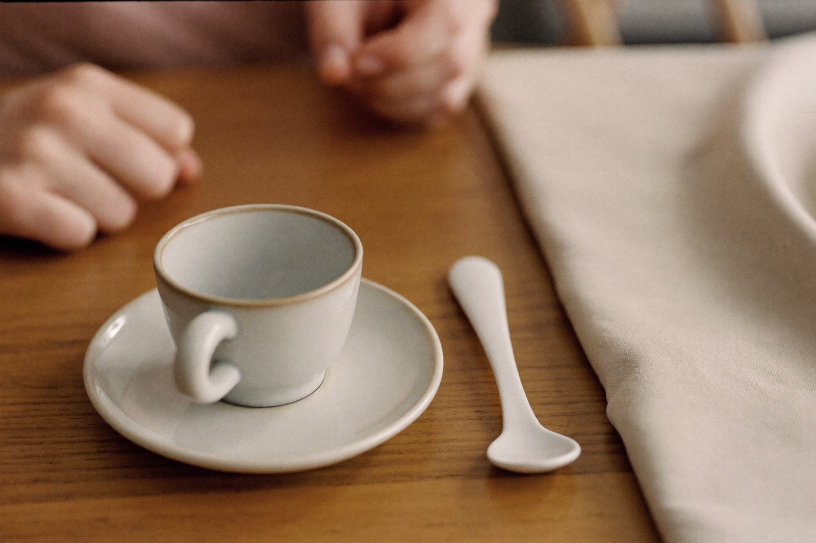 Taza y plato con una cuchara sobre una mesa de madera, con las manos de un adulto al fondo