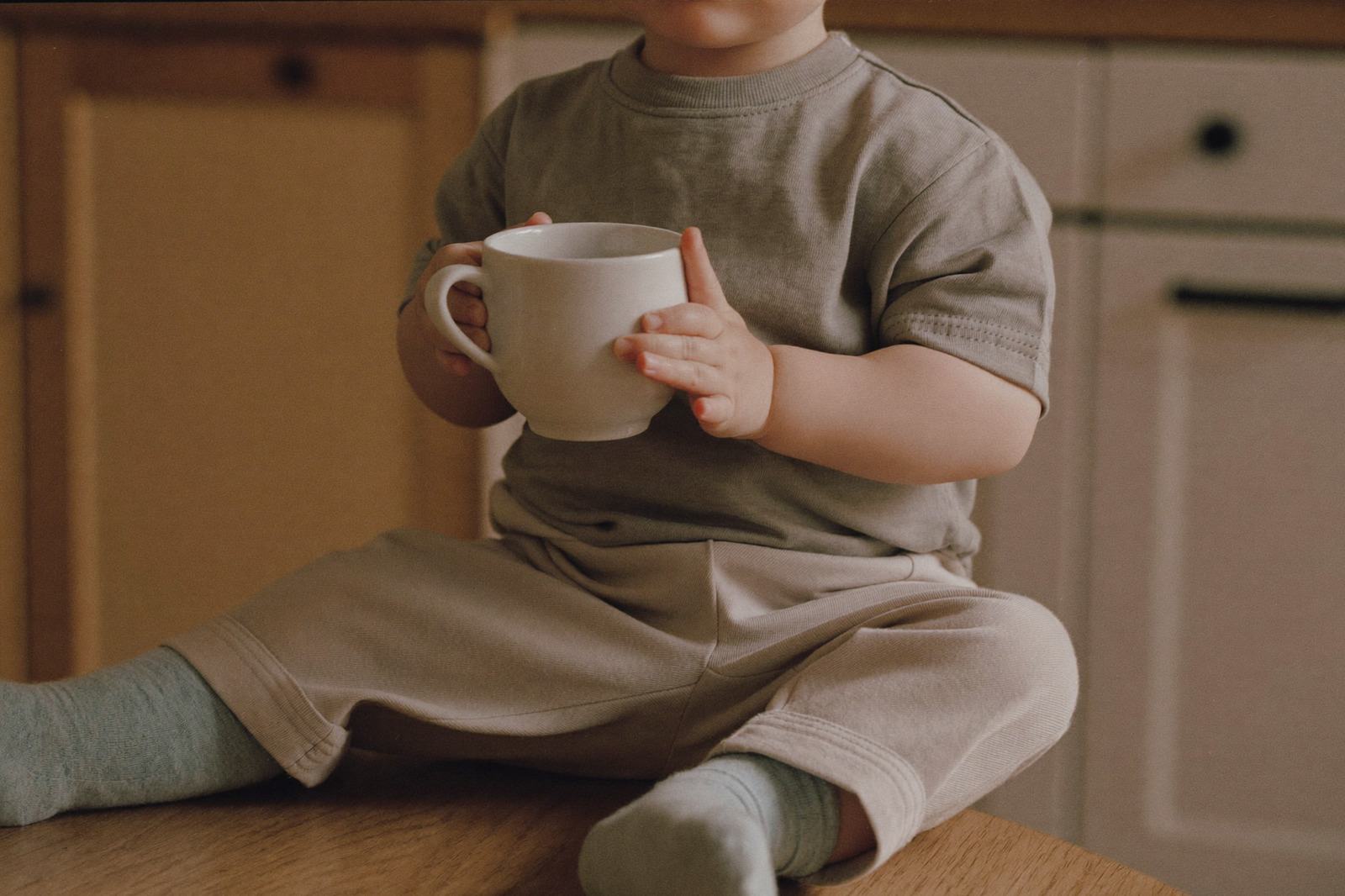 Niño pequeño sentado sobre una mesa de madera clara sosteniendo una taza con las dos manos