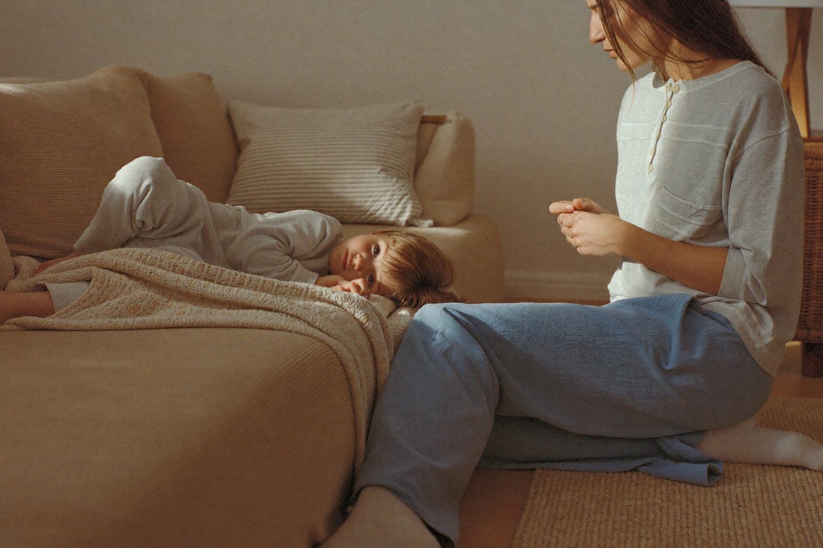 Niño pequeño recostado en una cama con una manta ligera y su mamá sentada en el piso, junto a él