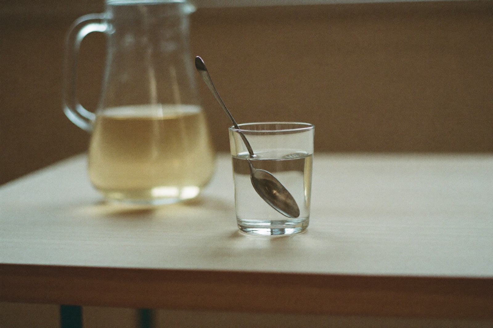 Vaso pequeño con líquido claro y una cuchara sobre una mesa junto a una jarra