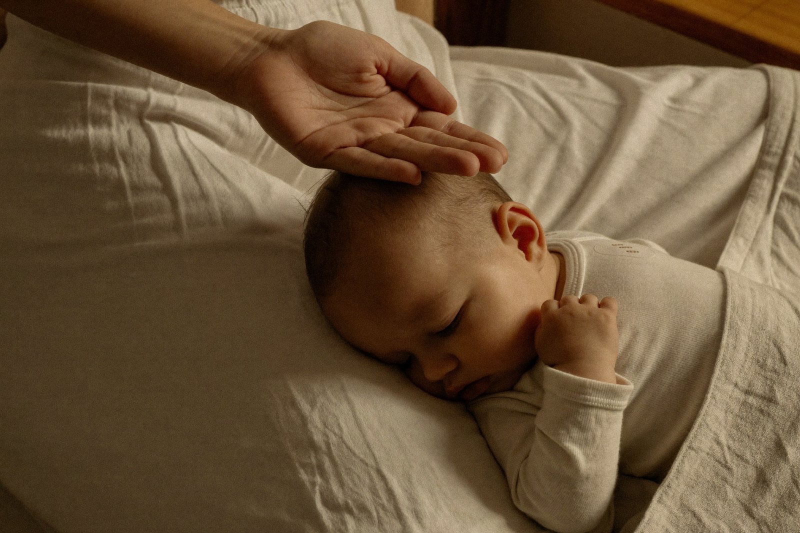 La mano de un adulto apoyada en la frente de un bebé dormido, con luz cálida y suave
