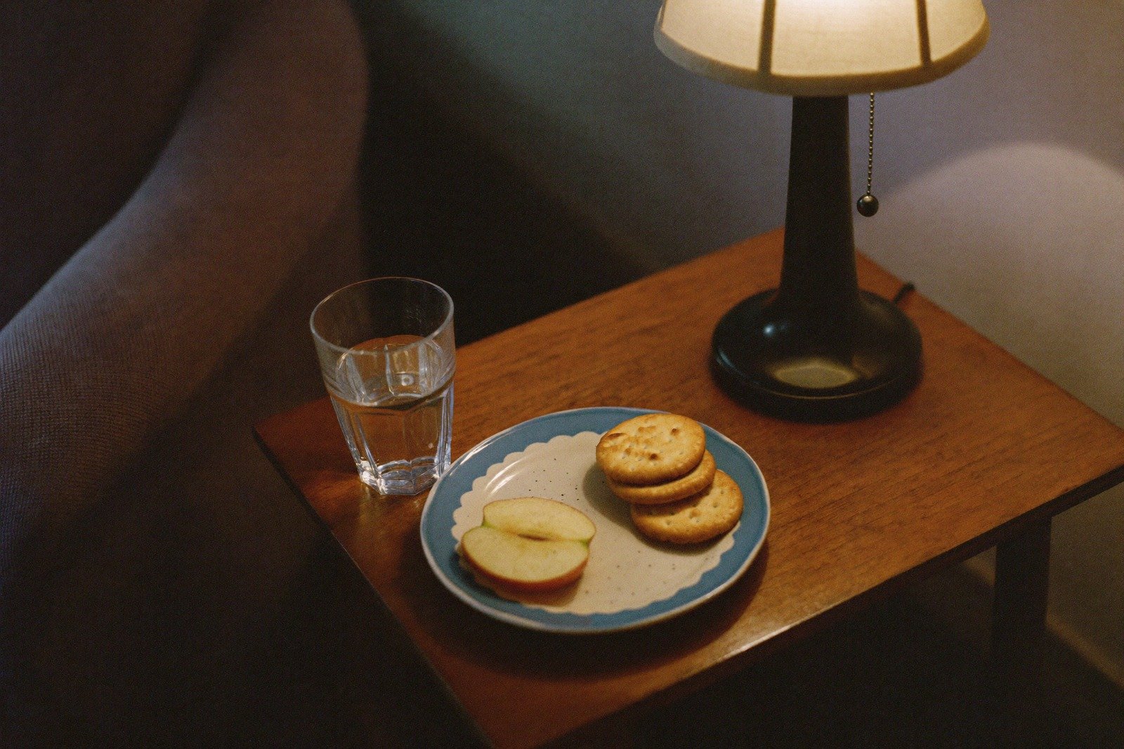 Vaso de agua y un plato con galletas y fruta sobre la mesa junto al sillón, con la lámpara encendida