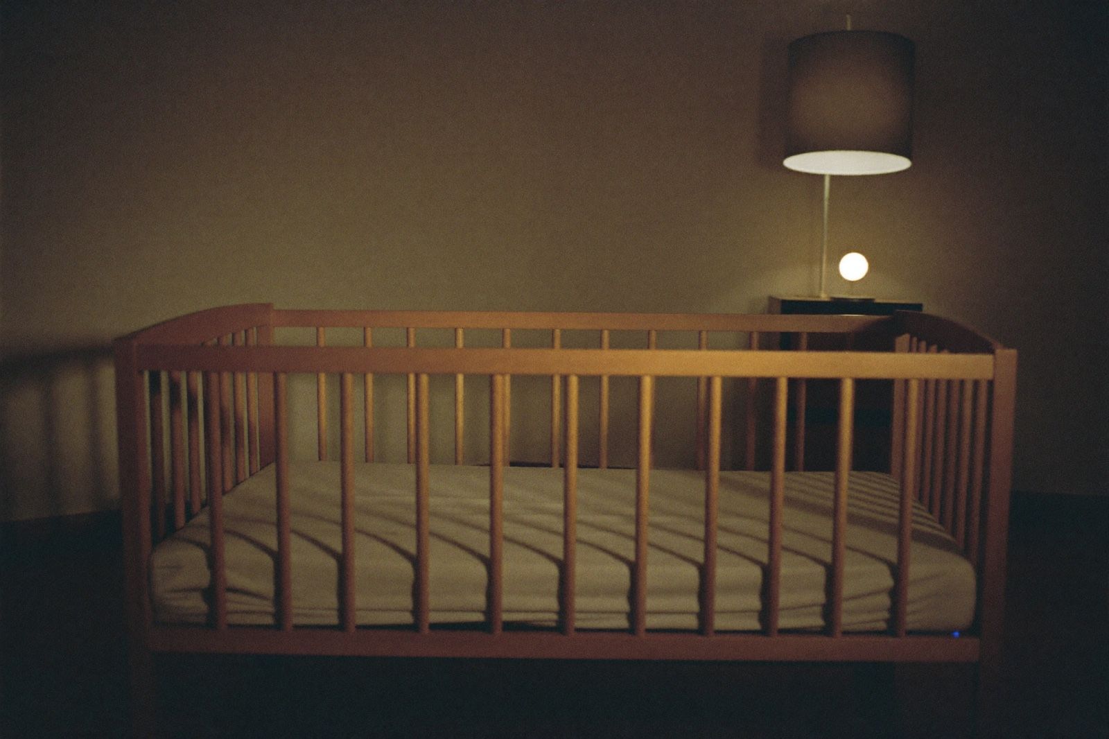 Cuna de madera vacía con sábana ajustada y nada más adentro, en una habitación en penumbra por la noche, con una lámpara cálida encendida en un rincón