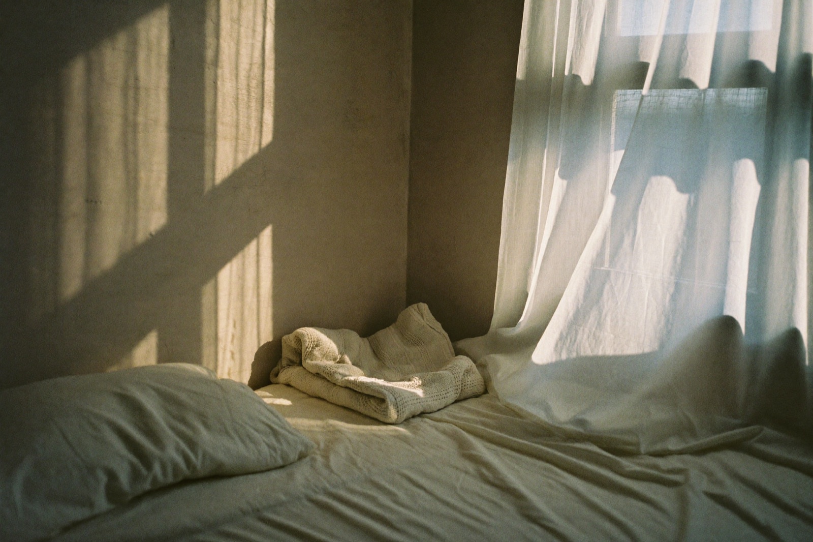 Cama con sábanas de lino claro y una manta tejida doblada al pie, con luz cálida de amanecer entrando por una cortina de gasa