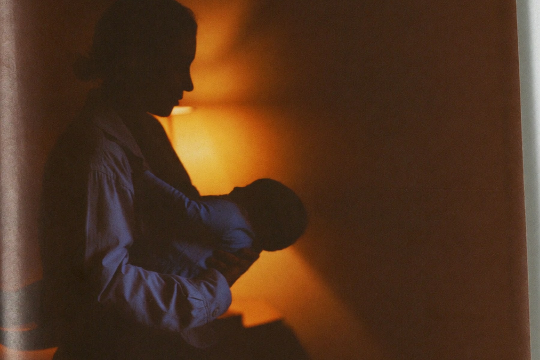La silueta de una madre de pie cargando a su bebé en una habitación a oscuras, recortada contra la luz ámbar de una lámpara