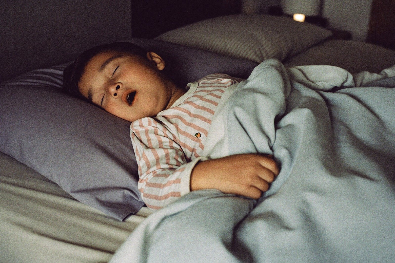 Niño pequeño dormido boca arriba en su cama con la boca entreabierta, en penumbra