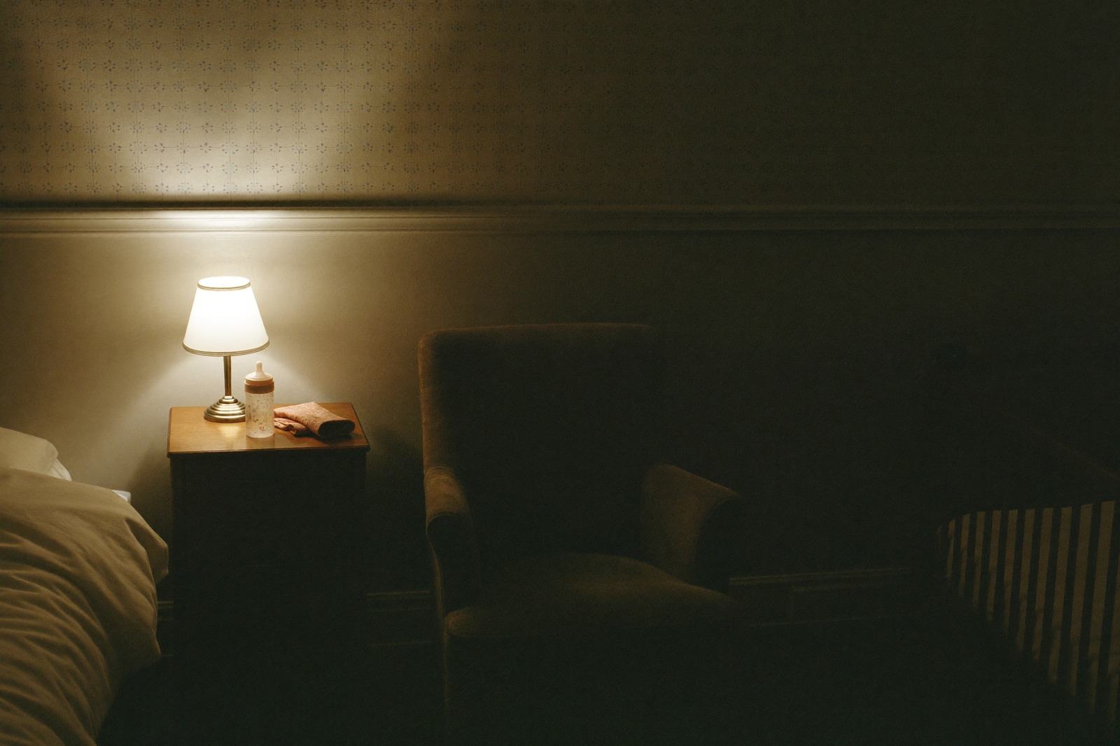 Habitación en penumbra con una lámpara tenue encendida, una mamila sobre el buró y un sillón junto a la cuna