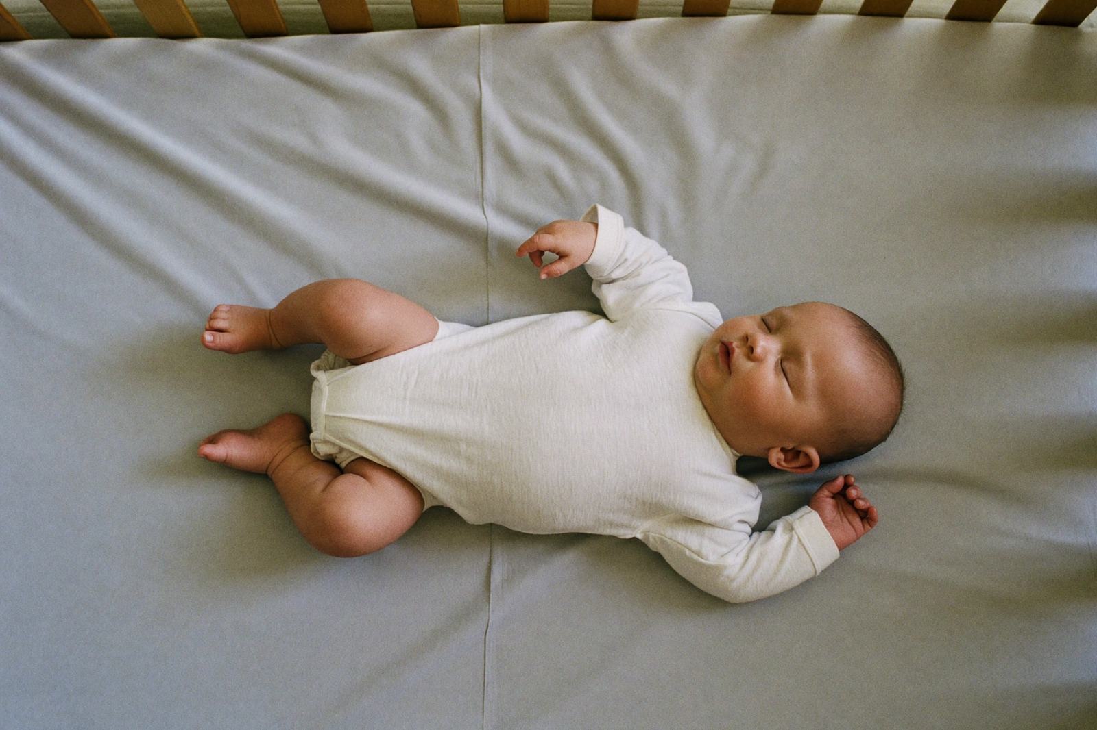 Bebé pequeño dormido boca arriba sobre la sábana ajustada de su cuna, con luz suave de día