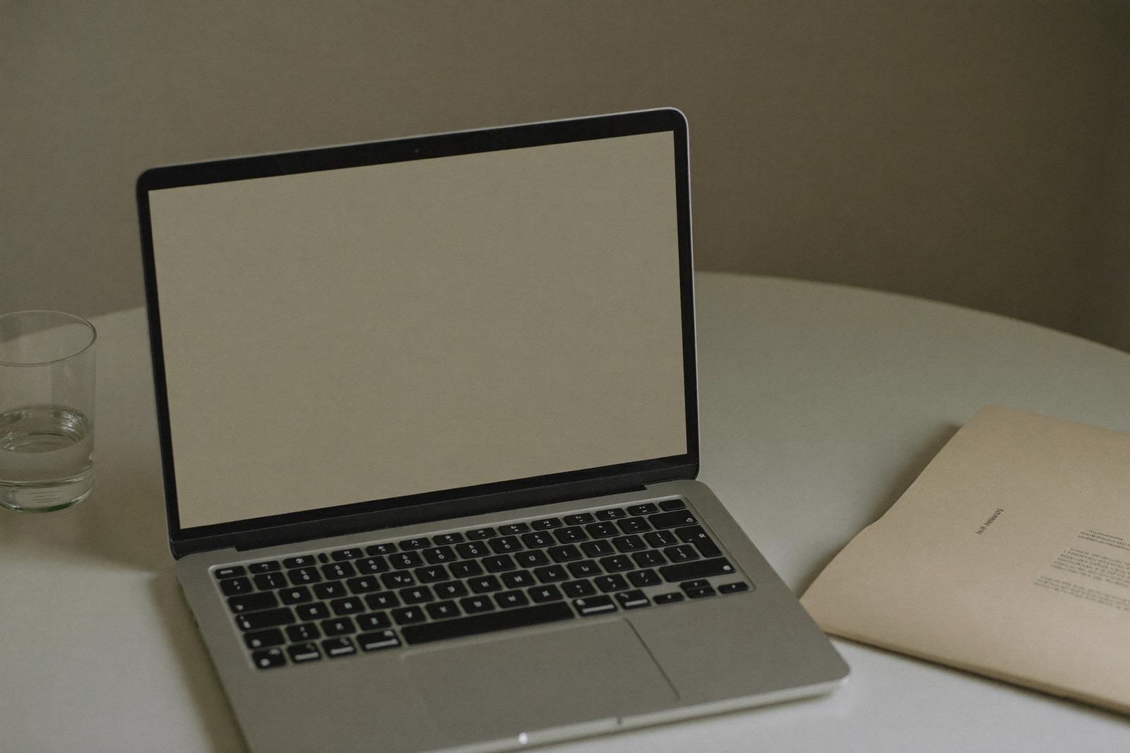 Laptop abierta sobre una mesa clara junto a una carpeta lisa y un vaso de agua