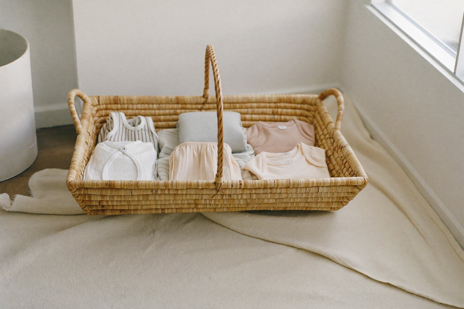 Canasta de mimbre con ropa de bebé doblada y una manta clara, sobre el piso de una habitación luminosa