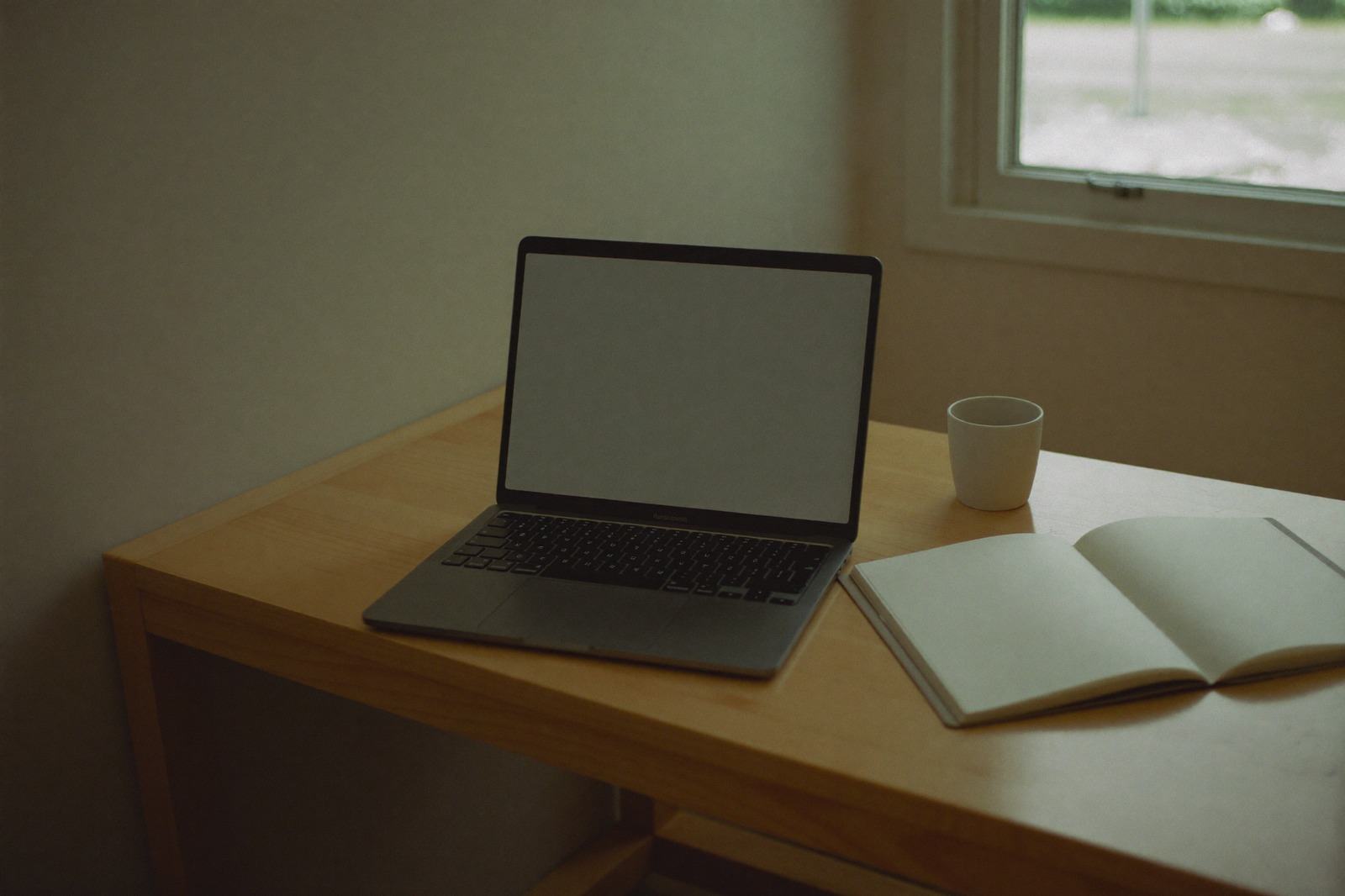 Escritorio claro con una laptop abierta, una carpeta lisa y una taza junto a la ventana