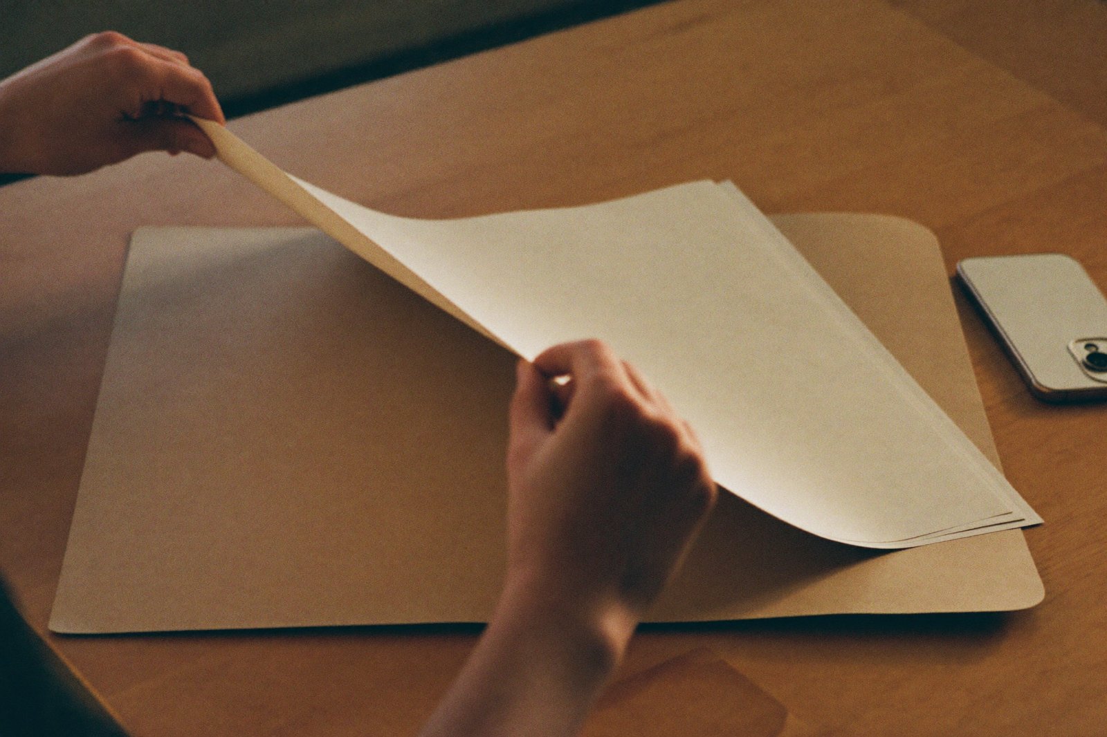 Unas manos guardando un documento dentro de una carpeta de papel sobre una mesa clara, con luz cálida de lado