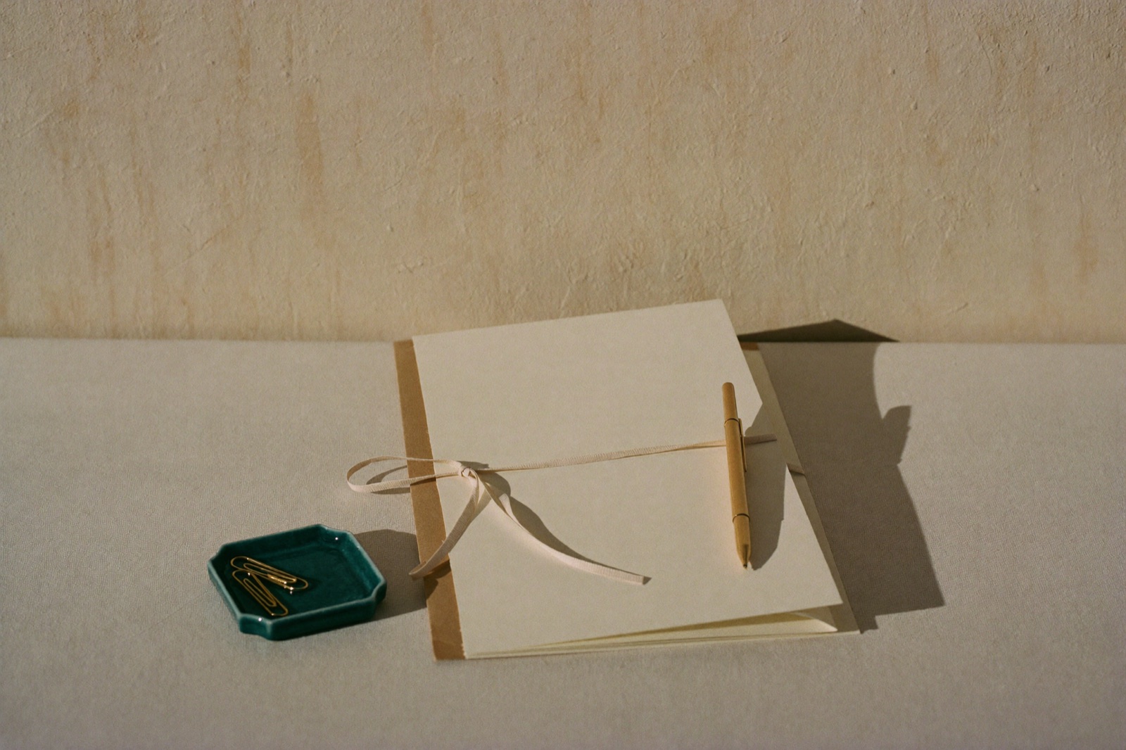 Fólder de papel kraft con hojas color crema atado con listón beige, una pluma de latón encima y un platito de cerámica azul verdoso con clips dorados, sobre una pared texturizada cálida