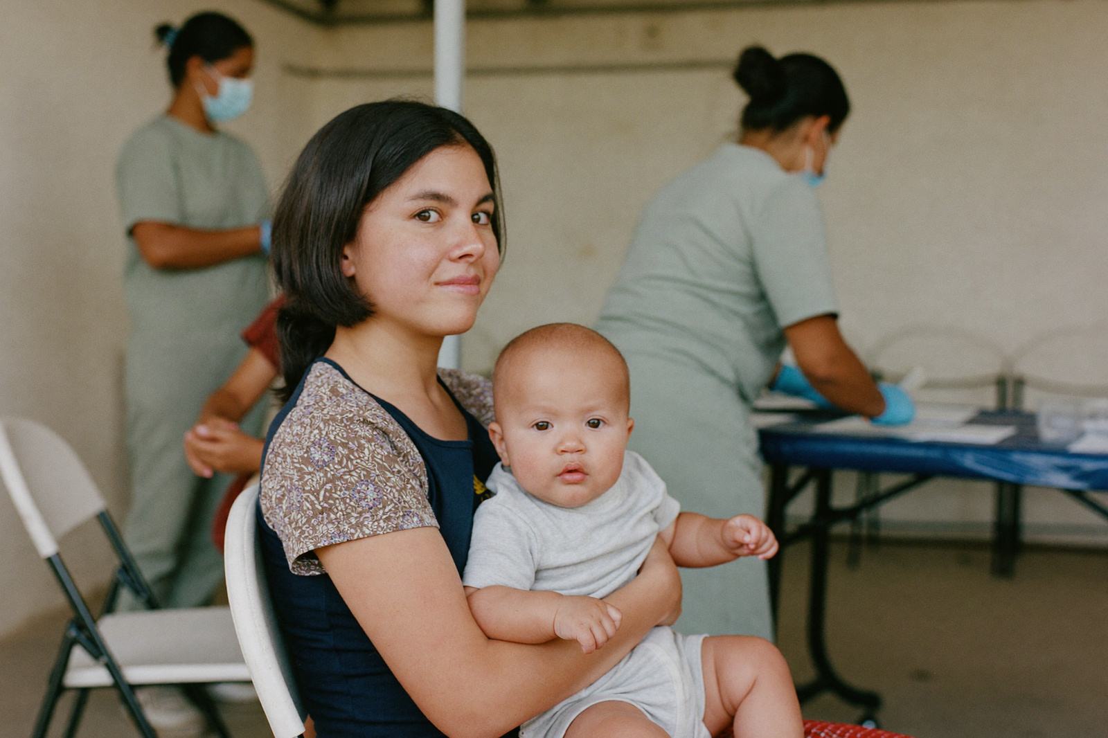 Madre con su bebé en brazos en un módulo de vacunación mientras el personal de salud atiende la mesa de registro al fondo