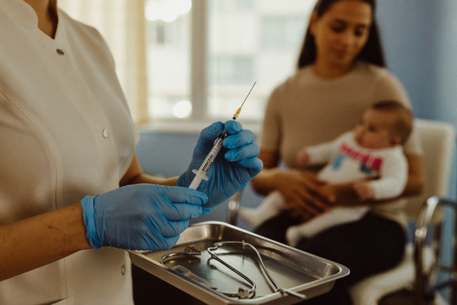 Una enfermera con guantes azules sostiene una jeringa preparada sobre una charola metálica; al fondo, una mamá con su bebé en brazos