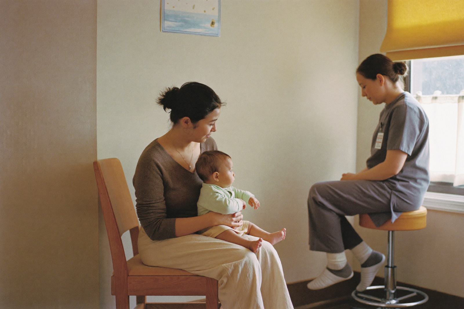 Madre sentada con su bebé en el regazo frente a la enfermera en un consultorio con luz de ventana, después de la consulta de vacunación