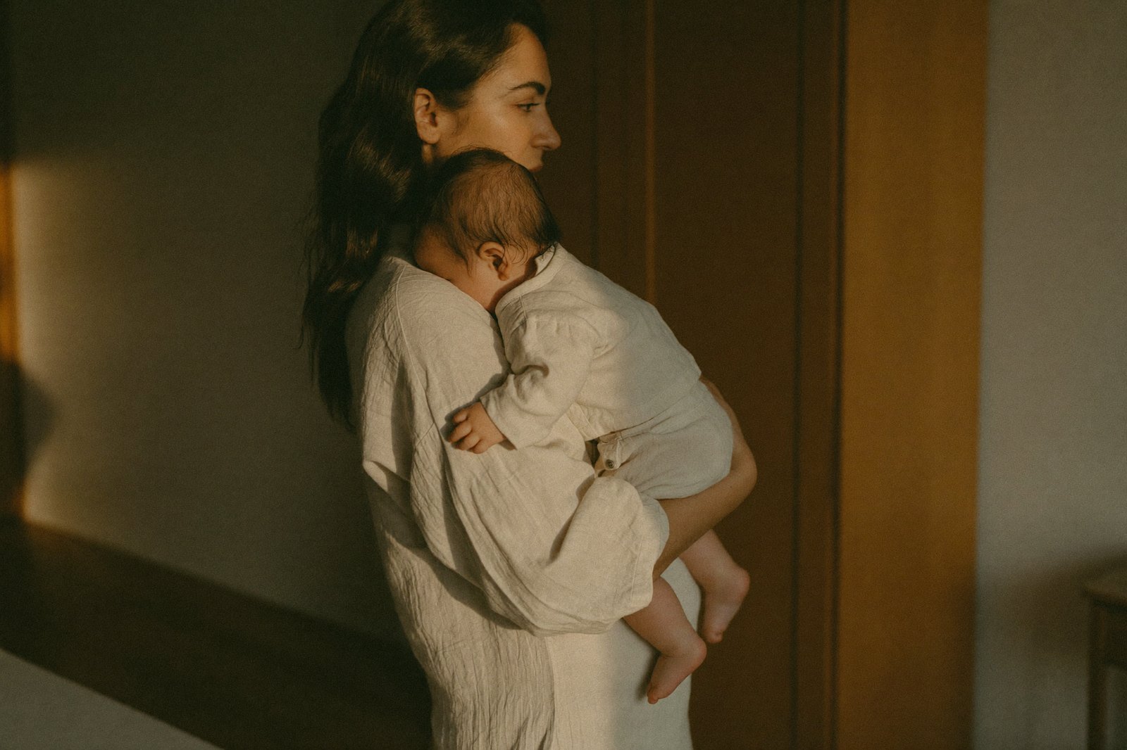 Una madre cargando a su bebé pegado al hombro, con la mano sosteniéndole la espalda, en una habitación con luz cálida
