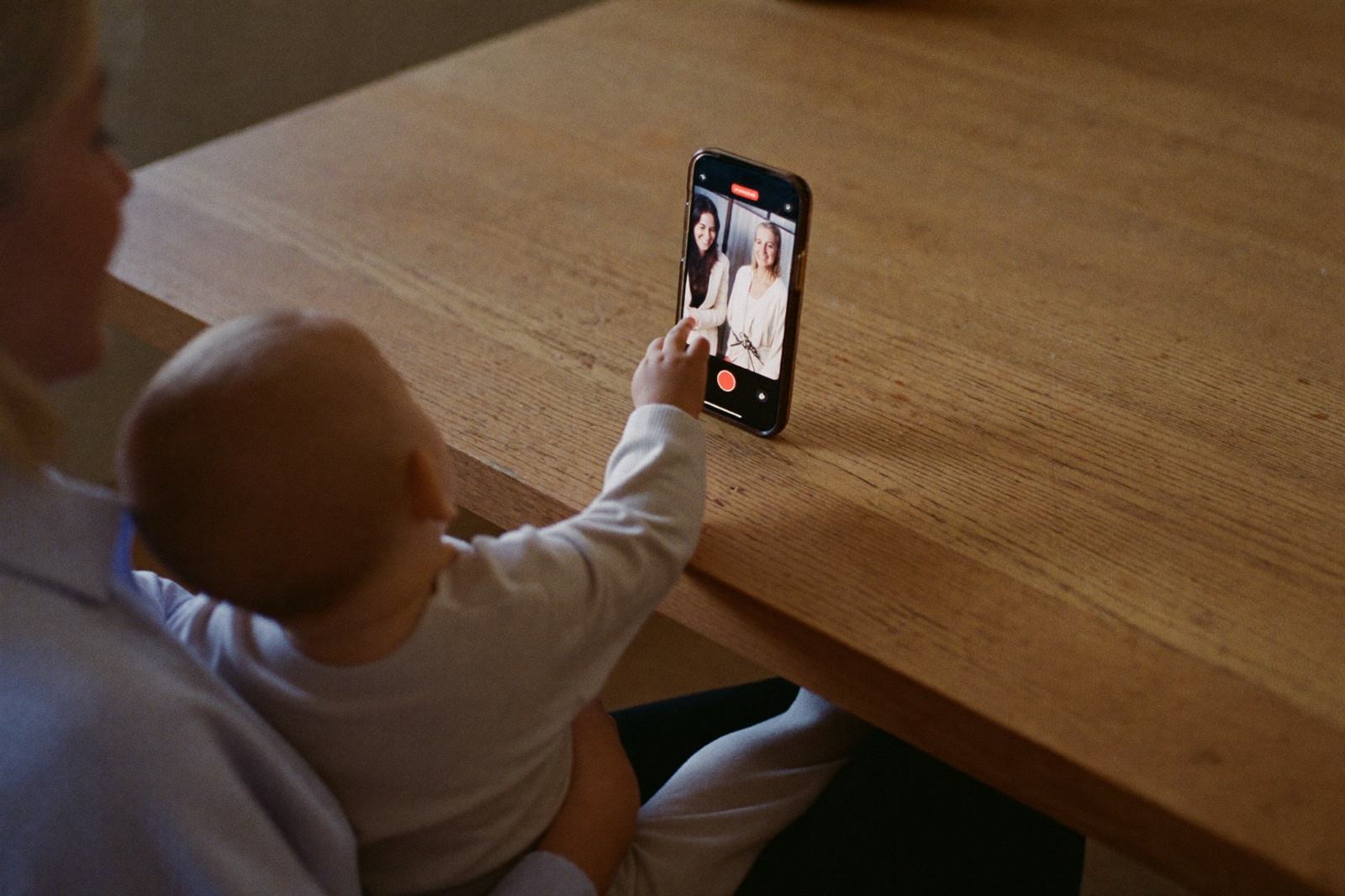 Un teléfono recargado en una mesa de cocina mostrando una videollamada con los abuelos, y un bebé sentado en las piernas de su mamá en primer plano estirando la mano hacia la pantalla
